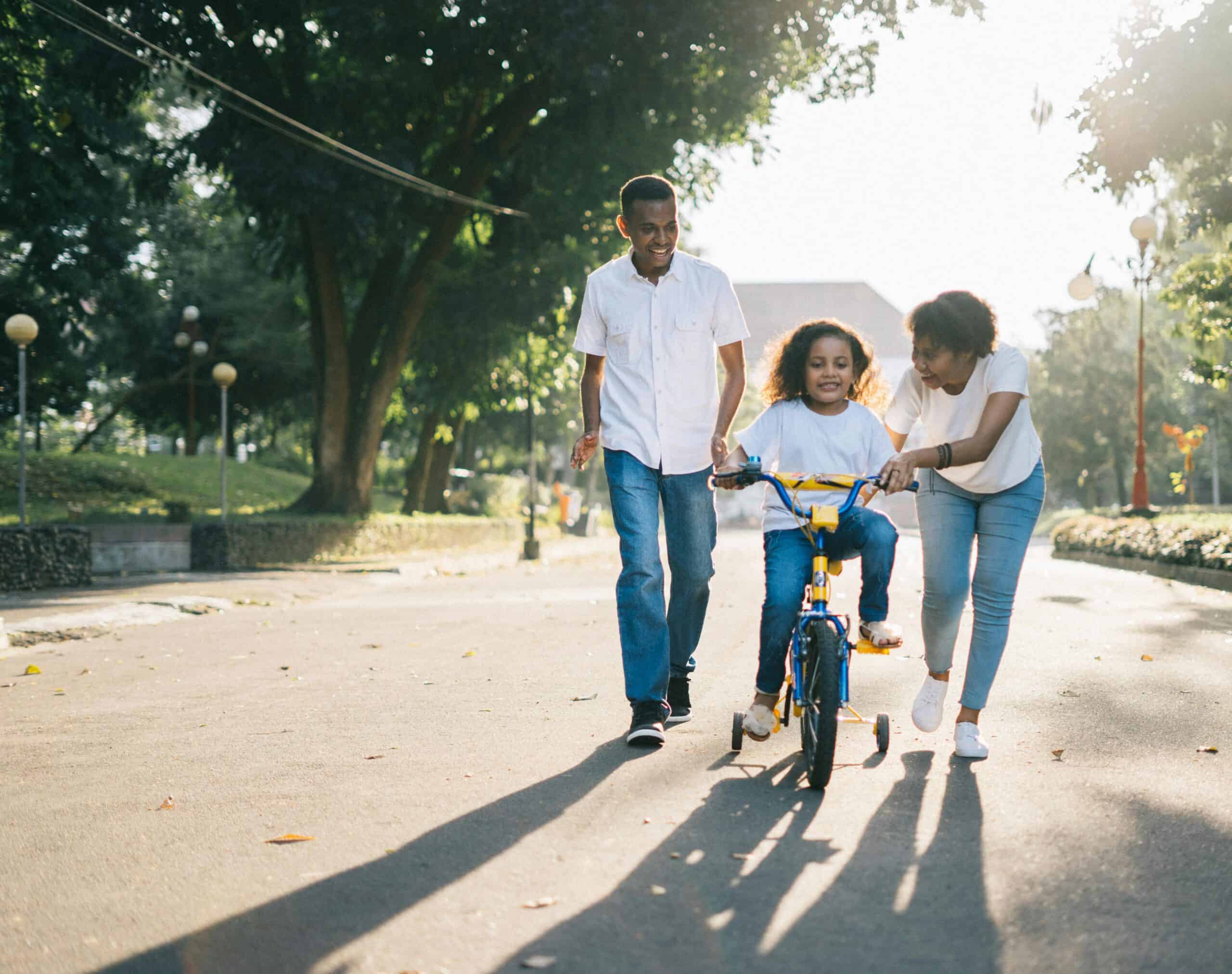 A family spending time together outdoors. A young child is riding a bicycle with training wheels, assisted by a smiling woman and accompanied by a man walking beside them.