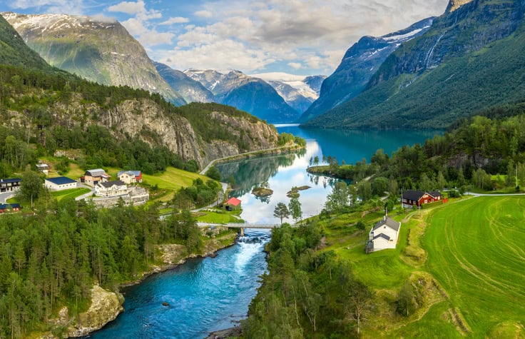 A breathtaking view of a serene valley featuring a turquoise river, traditional houses, and lush greenery framed by majestic snow-capped mountains under a partly cloudy sky.