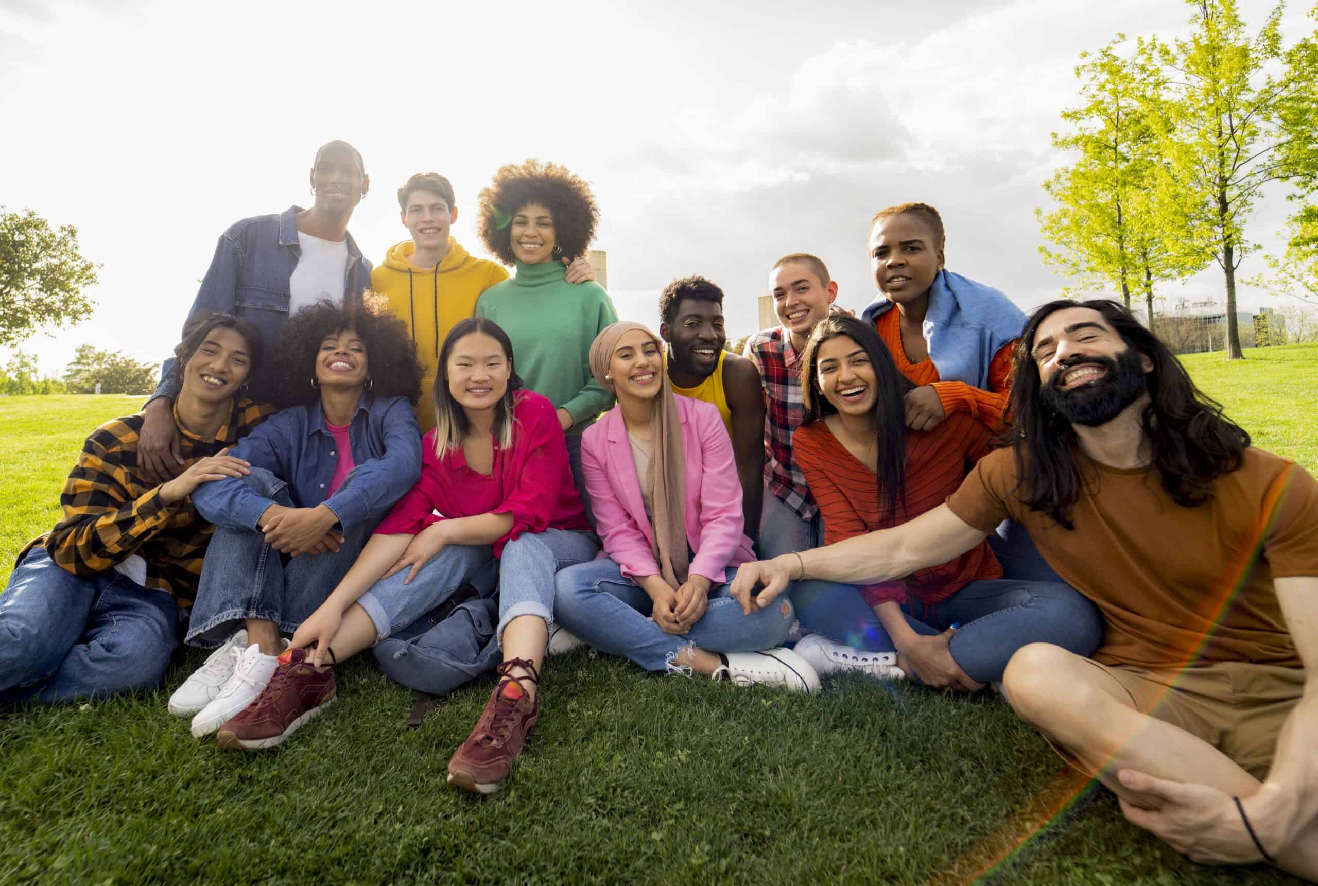 A diverse group of happy young people sitting together on a grassy field, smiling and laughing under a bright sky.