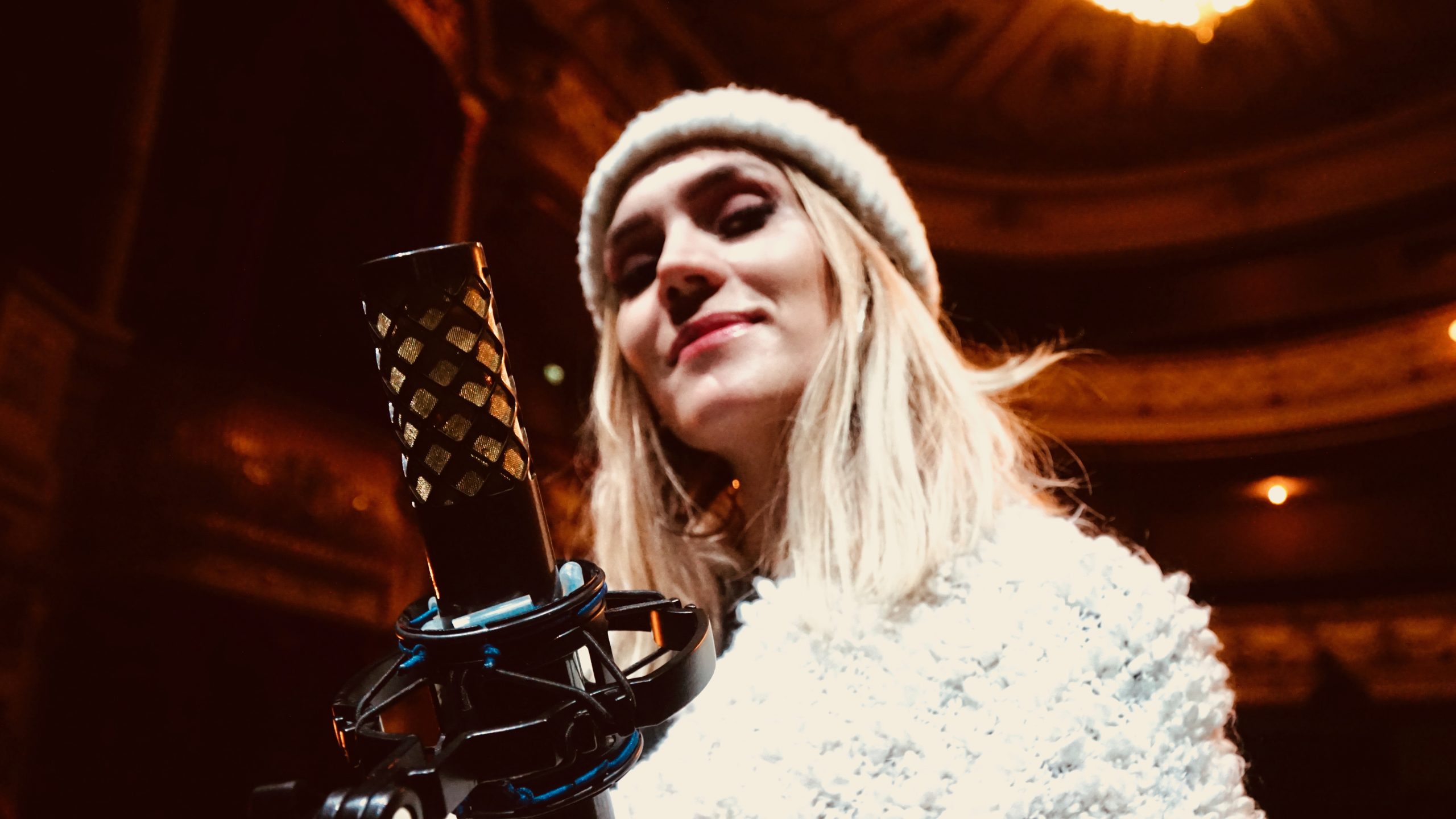 Female vocalist standing confidently behind a NU-100K microphone on stage in a warmly lit theater.