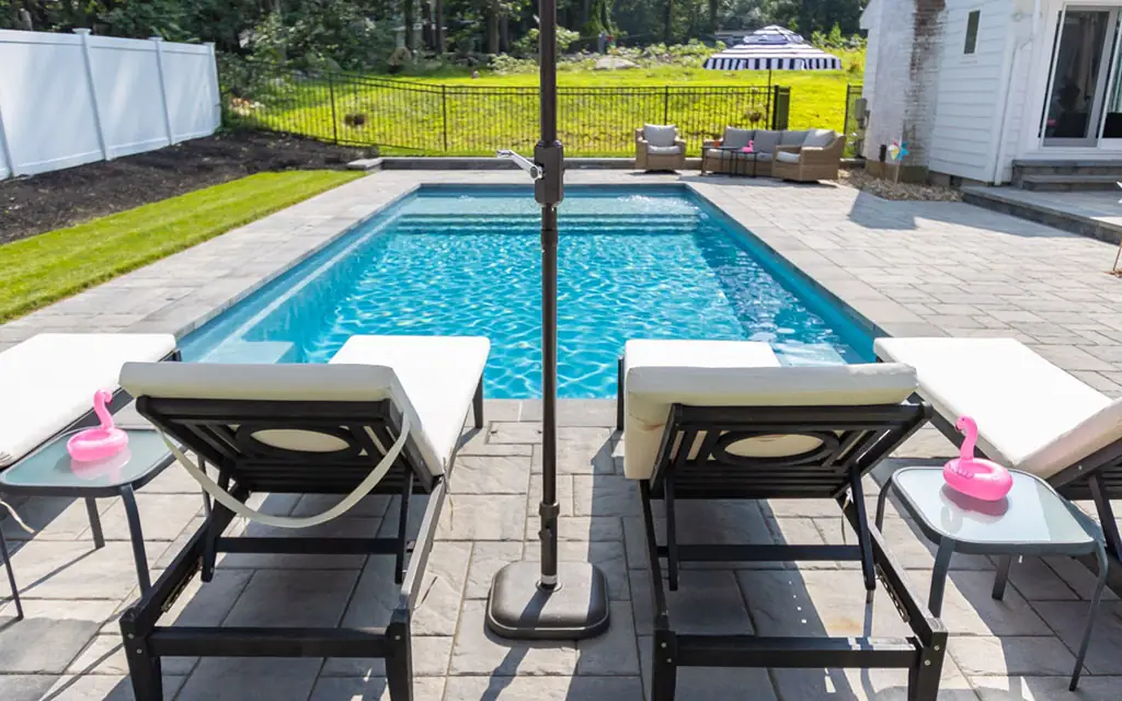 Two lounge chairs sit by a rectangular fiberglass pool with clear blue water. Small tables with pink flamingo cup holders are beside each chair. Lawn, fence, and patio furniture are visible—an inviting spot for your next pool project.