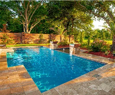 Rectangular outdoor swimming pool installation featuring clear blue water and stone tiles, nestled among lush landscaping and trees. Two small waterfalls flow at one end, with sunlight filtering through—the perfect setting for fiberglass or concrete swimming pools.