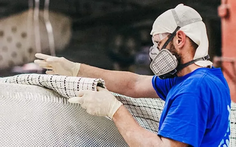 A person wearing a blue shirt, gloves, safety goggles, and a respirator mask handles a large sheet of industrial material in a workshop as part of concrete swimming pools or other pool projects.