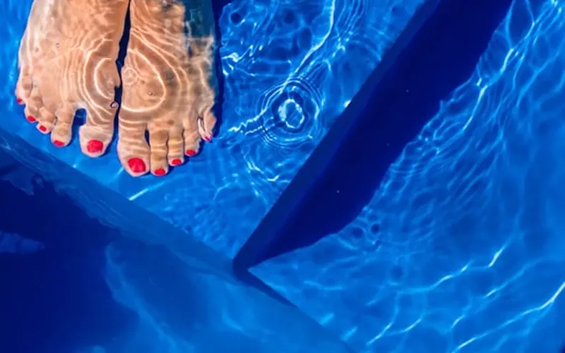 Bare feet with red-painted toenails rest on the edge of a fiberglass pool, sunlight reflecting on the water and creating wavy patterns on the skin and pool surface—perfect inspiration for your next swimming pool installation.