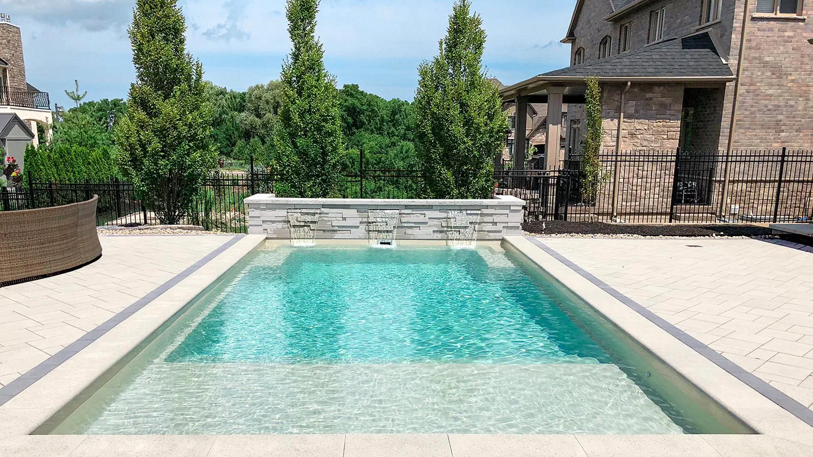 Rectangular backyard fiberglass pool with clear blue water, surrounded by light-colored stone tiles, adjacent to a brick house and green trees, under a partly cloudy sky—perfect for your next swimming pool installation.
