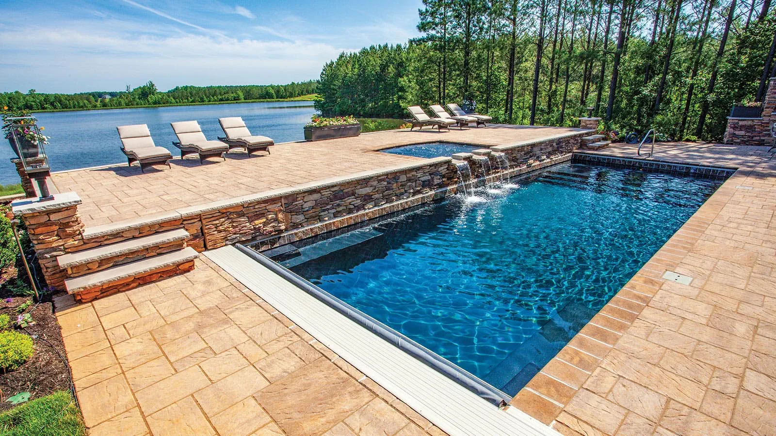 A rectangular concrete swimming pool with a waterfall feature and hot tub, surrounded by stone decking and lounge chairs, overlooks a lake and trees under a clear blue sky—perfectly crafted by an expert swimming pool contractor.
