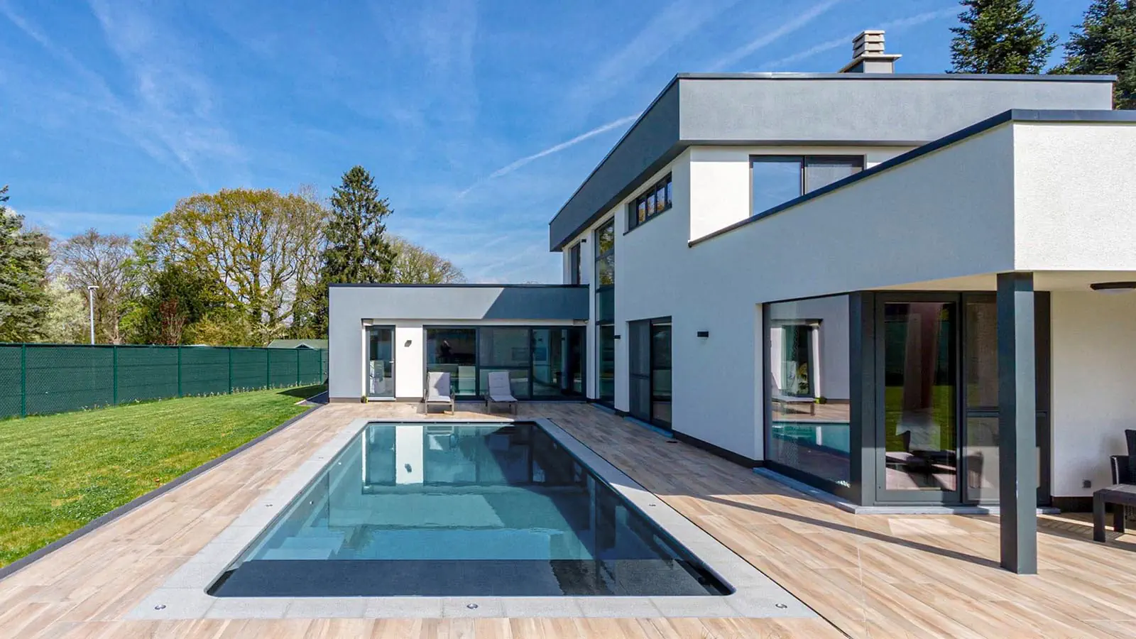 Modern two-story house with flat roof, large windows, and a sleek white and gray exterior. A concrete swimming pool installation is surrounded by a light wood deck, with green lawn and trees in the background under a clear blue sky.