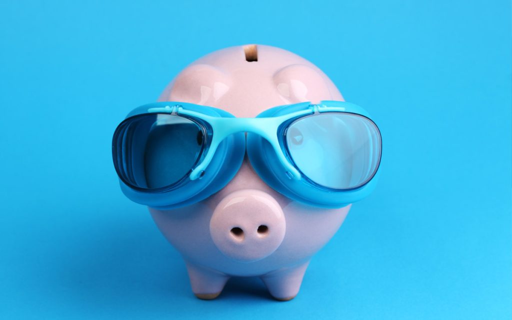 A pink piggy bank wearing blue swimming goggles stands against a solid blue background, ready to save for future fiberglass pools or exciting pool projects.