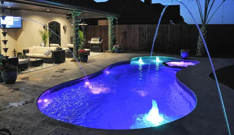 Custom swimming pool with deck jets and LED pool lighting at night.