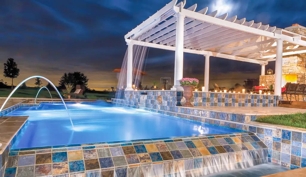 A modern backyard concrete swimming pool illuminated at night, featuring water features, a tiled infinity edge, and a pergola with string lights and seating beside a stone structure.