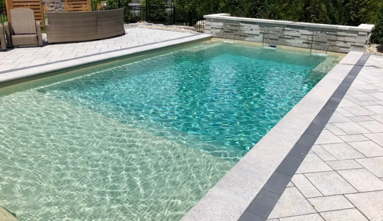 Rectangular outdoor swimming pool with clear turquoise water, surrounded by light-colored stone tiles and bordered by a low stone wall—an inspiring example of stylish fiberglass pools for your next swimming pool installation project. Patio furniture seen in the background.