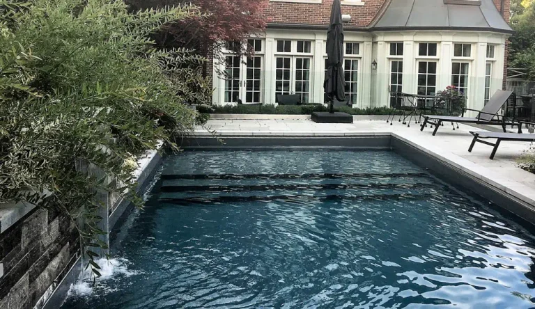 A modern rectangular fiberglass pool with steps, water feature, and clear blue water in a landscaped backyard; lounge chairs and an umbrella sit on the stone patio beside a large house. Built by a top swimming pool contractor.
