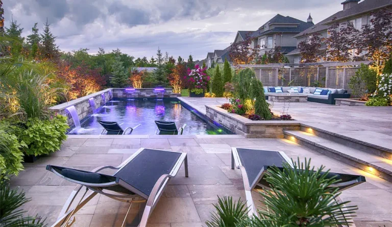 Modern backyard with a lighted fiberglass pool, surrounded by lush plants and lounge chairs, with a raised patio area featuring outdoor sofas and decorative landscaping—all expertly crafted by a professional swimming pool contractor.