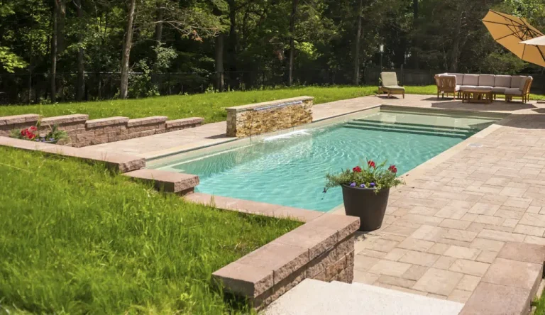 A rectangular fiberglass pool with stone edging, a small waterfall feature, potted flowers, and a patio area with outdoor seating and a large umbrella, all expertly crafted by a swimming pool contractor and surrounded by grass and trees.