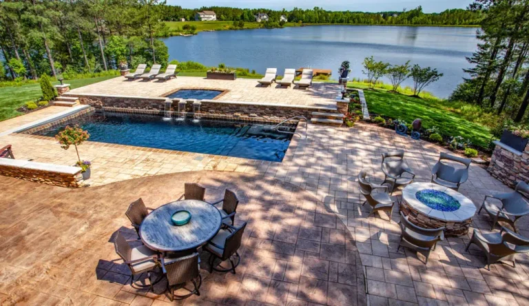 A spacious backyard patio features a dining table, fire pit with chairs, fiberglass pool with hot tub, lounge chairs, and a scenic view of a lake surrounded by trees and green grass—perfect inspiration for your next pool project.