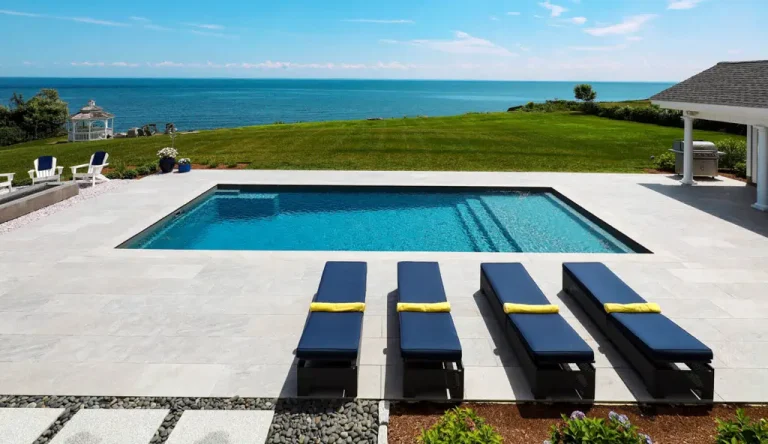 Luxury backyard featuring a rectangular concrete swimming pool installation with four navy lounge chairs with yellow towels, white stone patio, green lawn, and an ocean view under a blue sky.