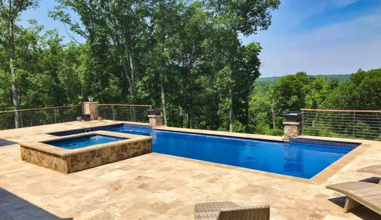 Rectangular concrete swimming pool with an attached hot tub on a stone patio, surrounded by lounge chairs and metal railing, overlooking a lush green forest under a clear blue sky—perfect for quality swimming pool installation.