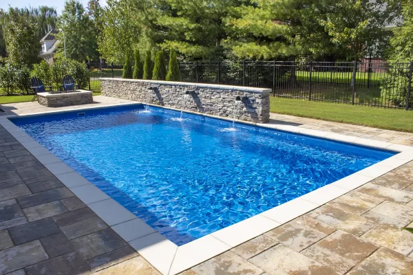 A rectangular inground swimming pool with clear blue water, built by a skilled swimming pool contractor, is surrounded by stone tiles and a water feature wall, set in a landscaped backyard with grass, trees, and a black metal fence.