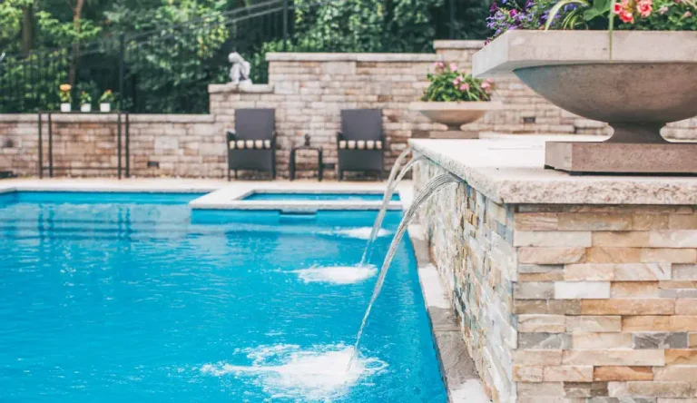 A backyard concrete swimming pool with clear blue water, stone wall fountains, lounge chairs, and potted flowers beside a brick wall and lush green foliage—perfect inspiration for your next pool project.
