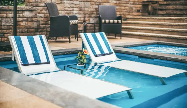 Two white lounge chairs with blue-and-white striped towels sit partially submerged in a pool—a stunning example of thoughtful pool projects by an expert swimming pool contractor. Wicker chairs and stone steps complete the inviting scene in the background.