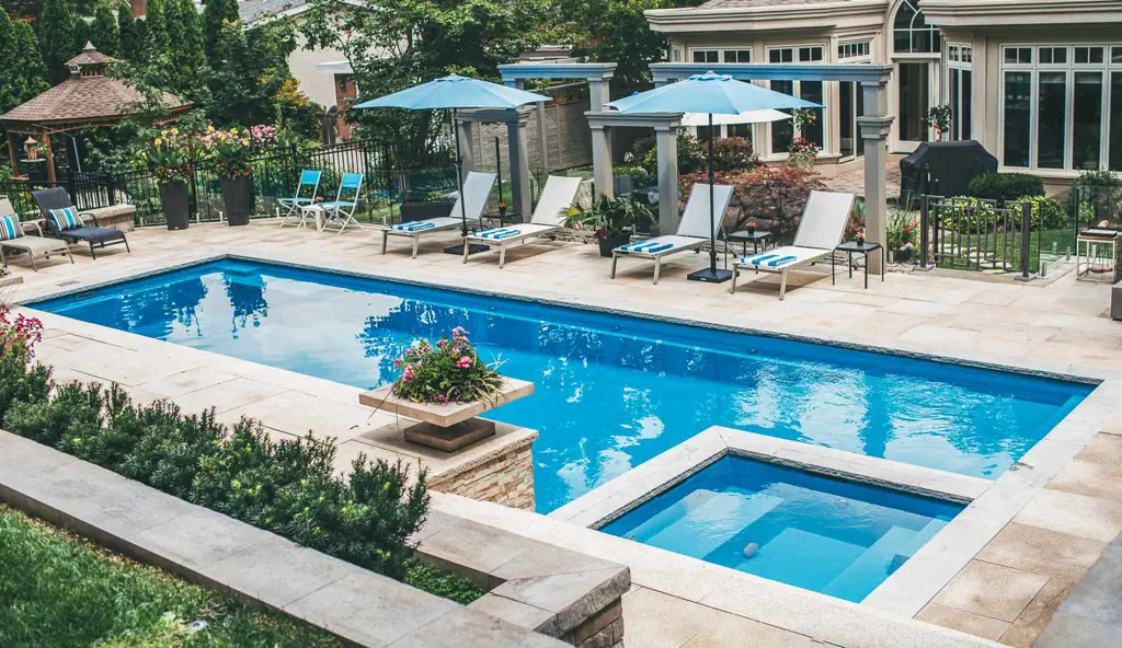 A backyard features a rectangular fiberglass pool with a small attached hot tub, lounge chairs with blue cushions, blue umbrellas, and lush gardens. Patio furniture, a gazebo, and expert swimming pool installation complete the inviting scene.