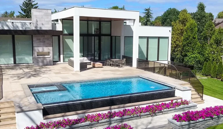 Modern house with large glass windows and patio, featuring a stunning concrete swimming pool with water fountains, surrounded by planters filled with pink flowers and a lush green lawn in the background.