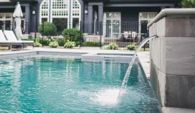 A modern backyard swimming pool installation features clear blue water and a stone fountain spout. Lounge chairs and greenery complement the dark-colored house with large windows, creating a stylish space for relaxing by concrete or fiberglass pools.