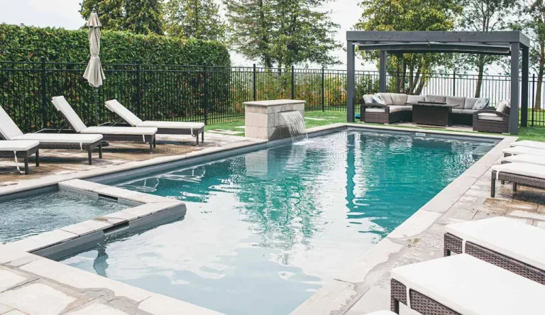 A modern backyard featuring a rectangular concrete swimming pool, surrounded by white lounge chairs, a large umbrella, and a shaded seating area. Lush trees and bushes line the fence—perfect inspiration for your next swimming pool installation.