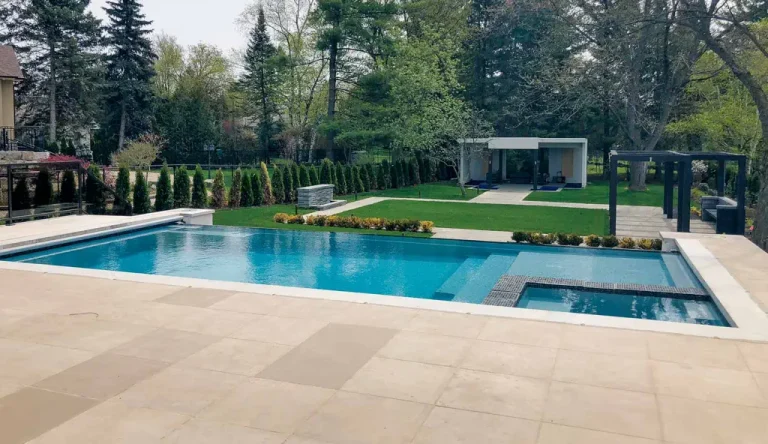 Modern backyard with a large rectangular fiberglass pool, hot tub, spacious patio, manicured lawn, hedges, and a small pool house or cabana in the background surrounded by trees—perfect for inspired pool projects.