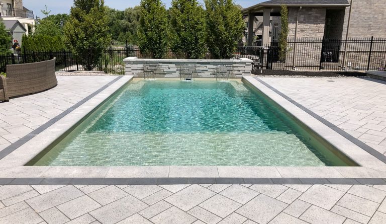 Rectangular concrete swimming pool with clear blue water, surrounded by light gray stone tiles. Trees and shrubs line the back, with a stone wall water feature at the far end—perfect inspiration for future pool projects.