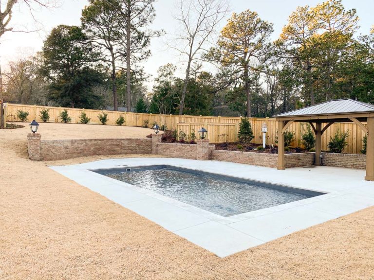 A backyard features a rectangular concrete swimming pool, surrounded by a patio and yellowed grass. Nearby are a wooden pergola, brick retaining wall, and privacy fence, all crafted with care by an expert swimming pool contractor.