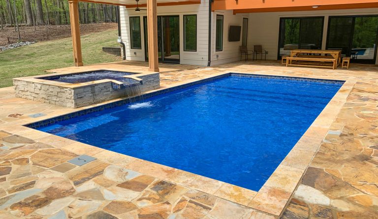 A backyard features a rectangular concrete swimming pool and attached hot tub, surrounded by stone tile decking. There’s a covered patio area with a wooden dining table and chairs, plus trees and grass in the background—perfect for relaxing after your swimming pool installation.