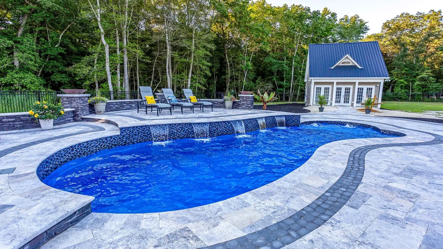 A backyard with a curvy, blue fiberglass pool featuring small waterfalls, surrounded by stone tiles. Lounge chairs with yellow pillows sit nearby. In the background, there’s a small house and trees.