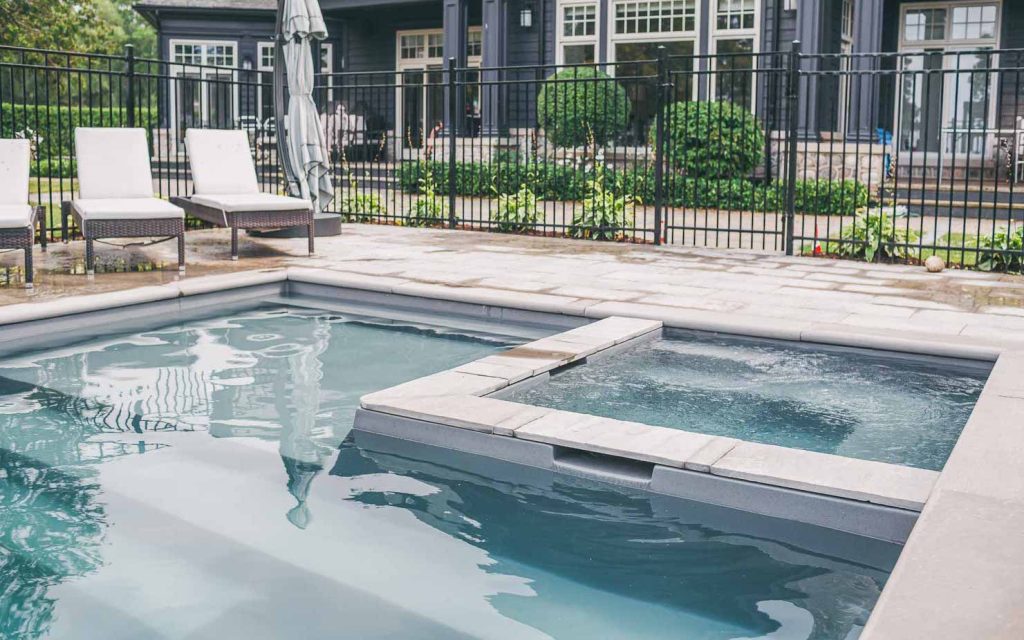 A backyard fiberglass pool with clear water features an attached hot tub separated by a stone ledge. Three white lounge chairs sit on a stone patio, with a black metal fence and house completing this stylish pool project.
