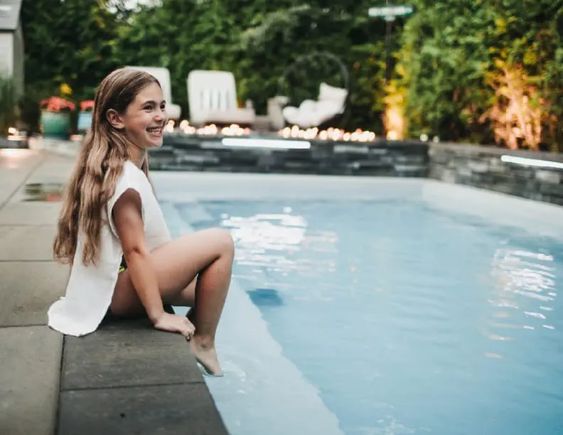A smiling young girl sits at the edge of a concrete swimming pool, dipping her feet in the water. The pool, installed by a skilled swimming pool contractor, is outdoors, surrounded by greenery and lit with evening lights.