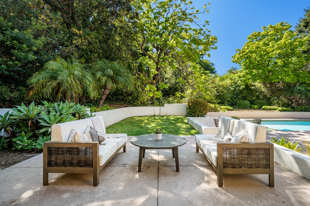 Two outdoor sofas with white cushions and a round table sit on a patio facing a lush green lawn and trees, with a beautiful fiberglass pool from recent pool projects visible to the right under a clear blue sky.