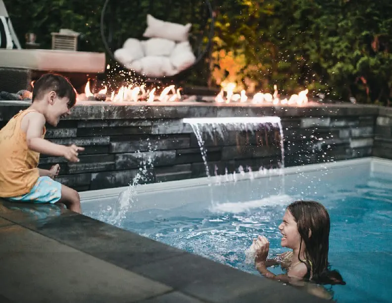 A young boy splashes water at a smiling woman in a backyard pool at dusk, where a stunning concrete swimming pool—crafted by an expert swimming pool contractor—features a fire display and waterfall, creating a warm, inviting atmosphere.