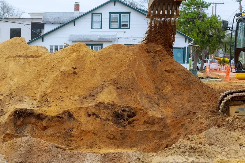 A large pile of sand in front of a house, with an excavator dumping more sand onto the pile at a construction site—perfect for pool projects like swimming pool installation. Trees and construction equipment appear in the background.