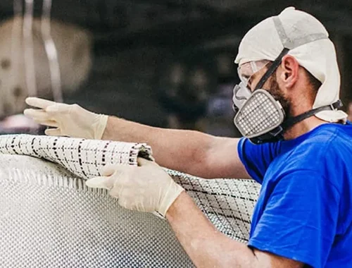 A man wearing a protective mask, gloves, and a hair cover works with a large sheet of material in an industrial setting, preparing components often used by swimming pool contractors for fiberglass pools and other pool projects.