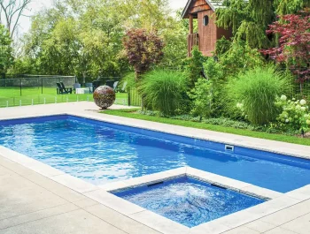A rectangular backyard swimming pool with an attached hot tub, expertly crafted by a swimming pool contractor, is surrounded by lush green landscaping, tall grasses, trees, and a wooden playhouse in the background.