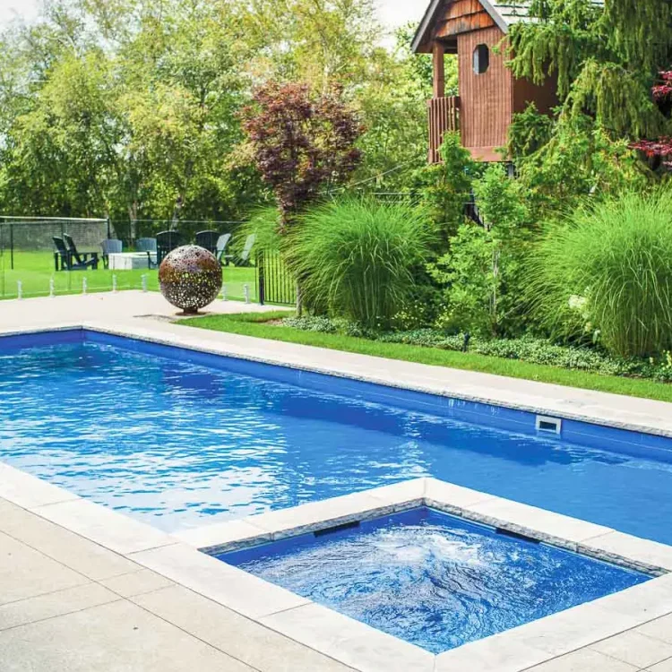 A rectangular backyard swimming pool with an attached hot tub, expertly crafted by a swimming pool contractor, is surrounded by lush green landscaping, tall grasses, trees, and a wooden playhouse in the background.