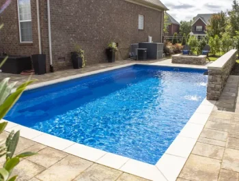 A rectangular backyard fiberglass pool with clear blue water is surrounded by stone tiles, potted plants, and outdoor furniture. A brick house sits in the background on a sunny day—ideal inspiration for your next swimming pool contractor project.