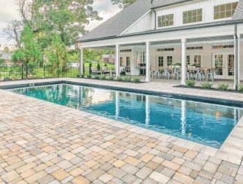 A modern backyard with a rectangular fiberglass pool, surrounded by a stone patio, outdoor seating, and a large house with tall windows, columns, and a covered porch—a stunning example of expert pool projects.