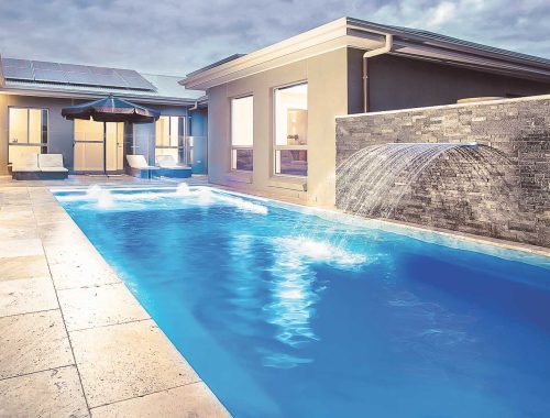 Modern backyard with a rectangular fiberglass pool installed beside the house, stone-tiled deck, water feature with arched spout, lounge chairs, and umbrella—all lit with soft outdoor lighting at dusk by a skilled swimming pool contractor.