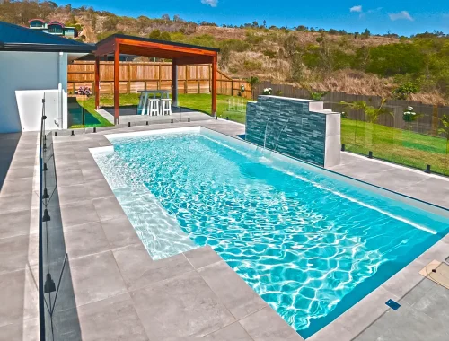 Modern backyard with a rectangular concrete swimming pool, waterfall feature, gray tile deck, glass fence, covered patio with seating, and green lawn surrounded by a wooden fence and trees. Perfect for enjoying expert swimming pool installation.