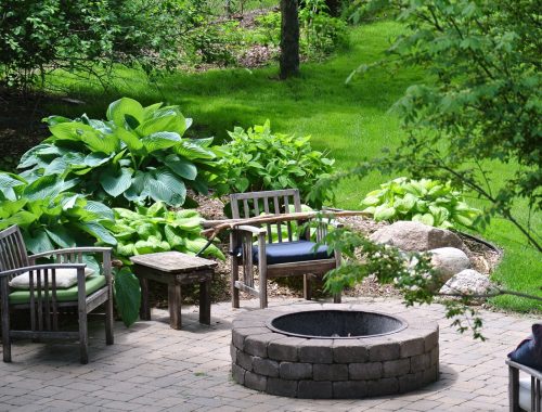 A stone fire pit is surrounded by wooden chairs with cushions and a small table, set on a patio. Lush green hostas and trees fill the garden, while a swimming pool contractor can help add fiberglass pools to enhance this peaceful outdoor space.
