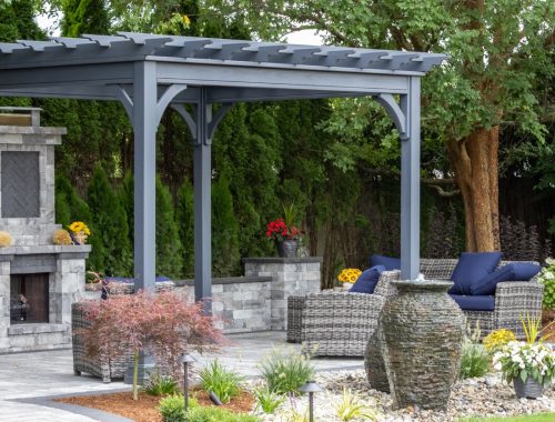 A modern backyard patio with a gray pergola, wicker furniture with blue cushions, an outdoor fireplace, large stone planters, lush greenery, and space ideal for new pool projects by a trusted swimming pool contractor.