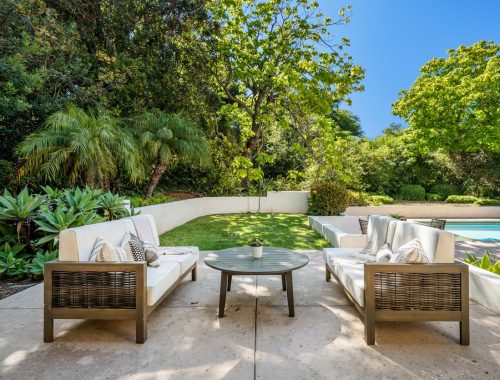 Two outdoor sofas with white cushions and a round table sit on a patio facing a lush green lawn and trees, with a beautiful fiberglass pool from recent pool projects visible to the right under a clear blue sky.
