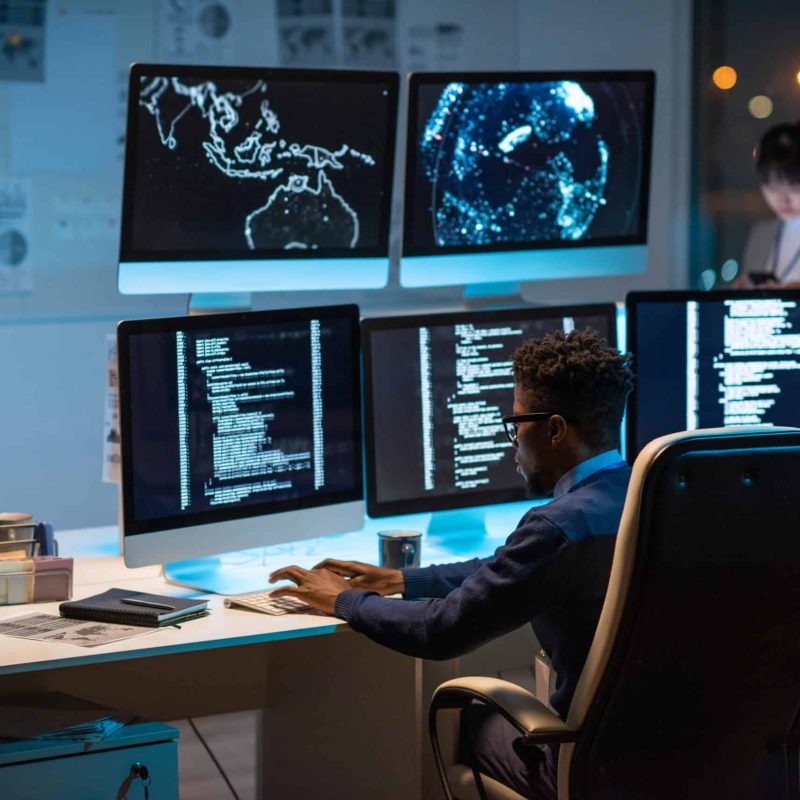 Young contemporary cyber security manager typing while sitting by desk in front of computer monitors