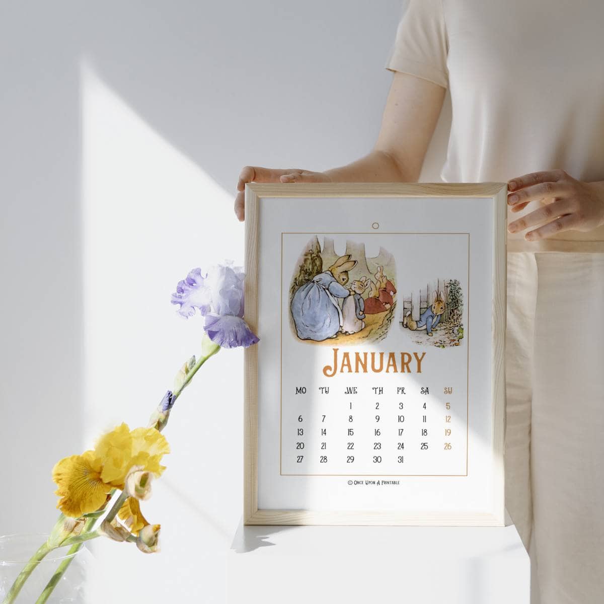 Peter Rabbit illustrated January calendar in a frame with a woman holding it next to Iris's in a vase.